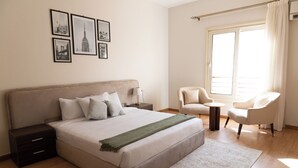 A bedroom with a large bed, bedside tables, a chair, a small table, and a window with curtains. - The Seven Six Near Waterway 5A - 3, THE SEVEN SIX near WATERWAY 5A MALL By ELEGANT (New Cairo City)