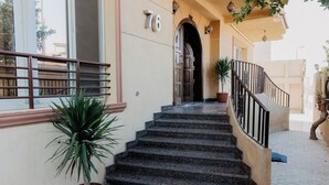 A staircase leading to a door with the number 76, flanked by potted plants and a railing. - The Seven Six Near Waterway 5A - 4, THE SEVEN SIX near WATERWAY 5A MALL By ELEGANT (New Cairo City)