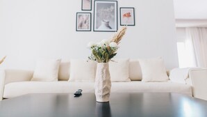 A living room with a white sofa, a coffee table with a vase of flowers, and framed pictures on the wall. - The Seven Six Near Waterway 5A - 4, THE SEVEN SIX near WATERWAY 5A MALL By ELEGANT (New Cairo City)