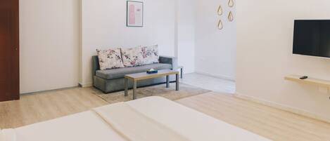 A modern hotel room with a large bed, a grey sofa with floral cushions, a small table, and a TV mounted on the wall.