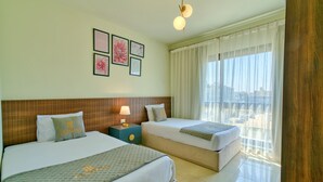 A hotel room with two beds, a nightstand with a lamp, framed artwork on the wall, and a large window with curtains.