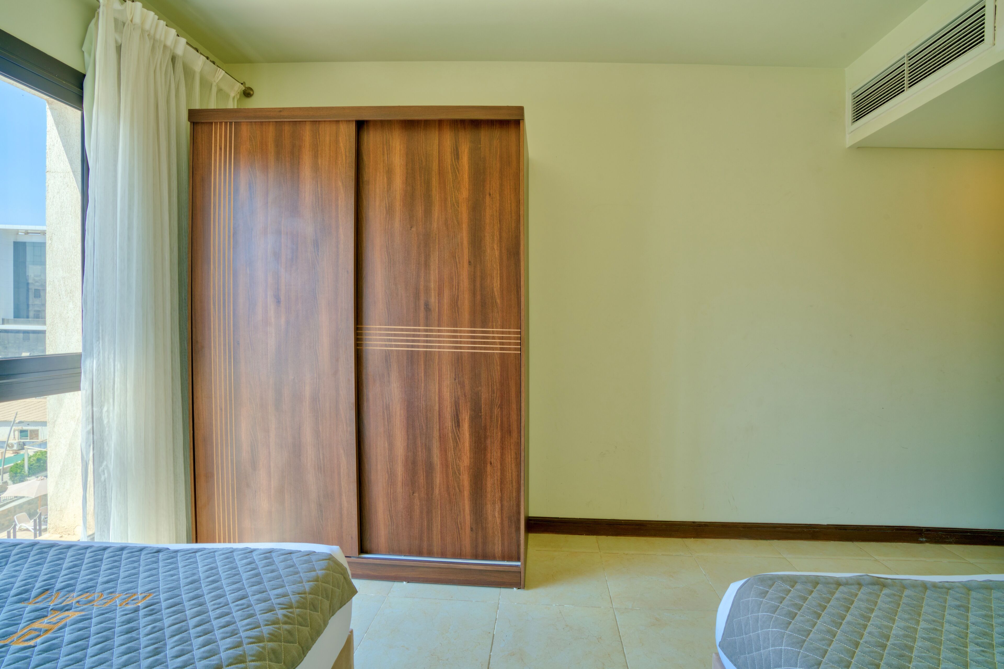 A hotel room with a large wooden wardrobe, a bed with a quilted cover, and a window with curtains.