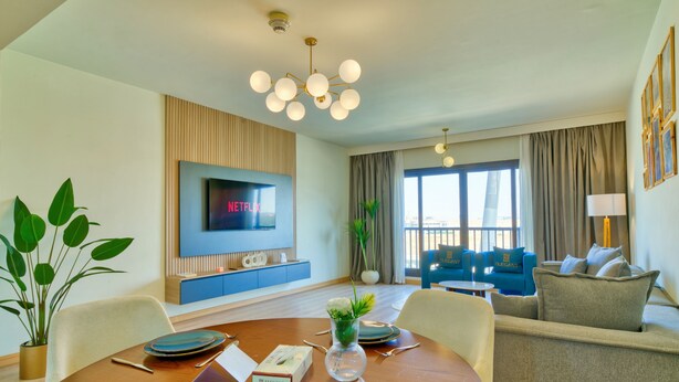 A modern hotel room with a dining area, a living area with a sofa, a TV mounted on the wall, and a large window with curtains.
