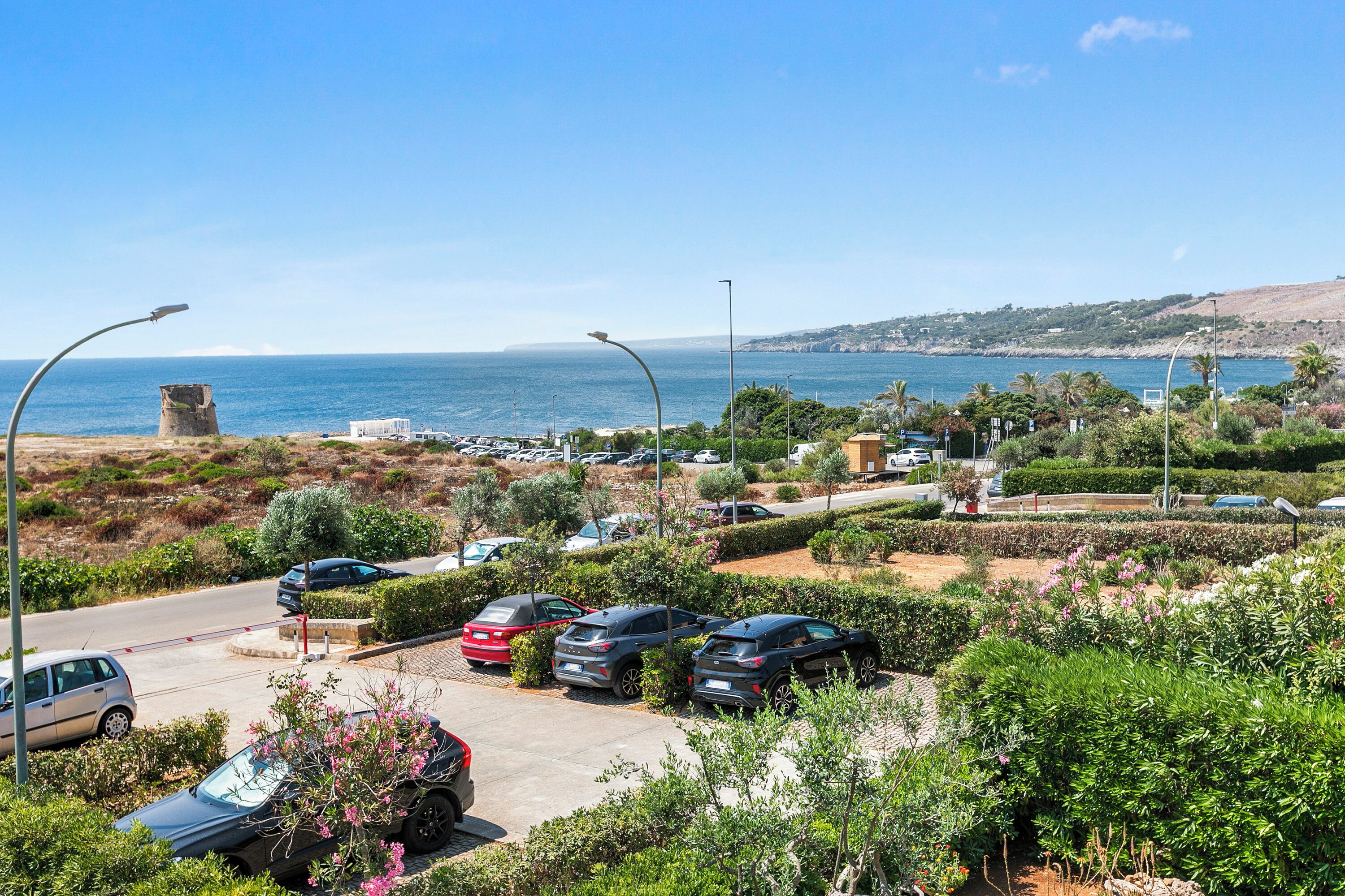 A coastal parking area with cars, greenery, and a clear view of the sea.