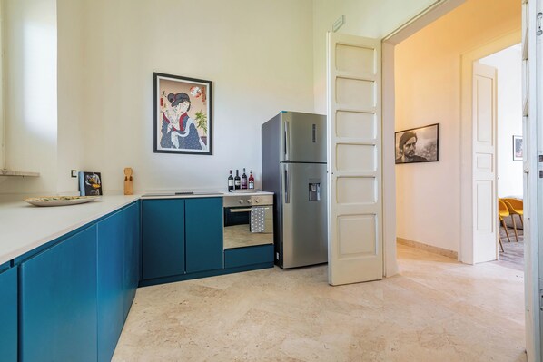 A modern kitchen with blue cabinets, a stainless steel refrigerator, and a small oven. There are framed pictures on the wall and a door leading to another room.