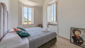 Ein Schlafzimmer mit einem Bett, zwei Fenstern, einem Sessel und einem Bild an der Wand.
