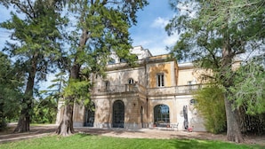 Ein historisches Gebäude mit großen Bäumen im Vordergrund, einem gepflasterten Innenhof und einem Balkon mit Gartenmöbeln.