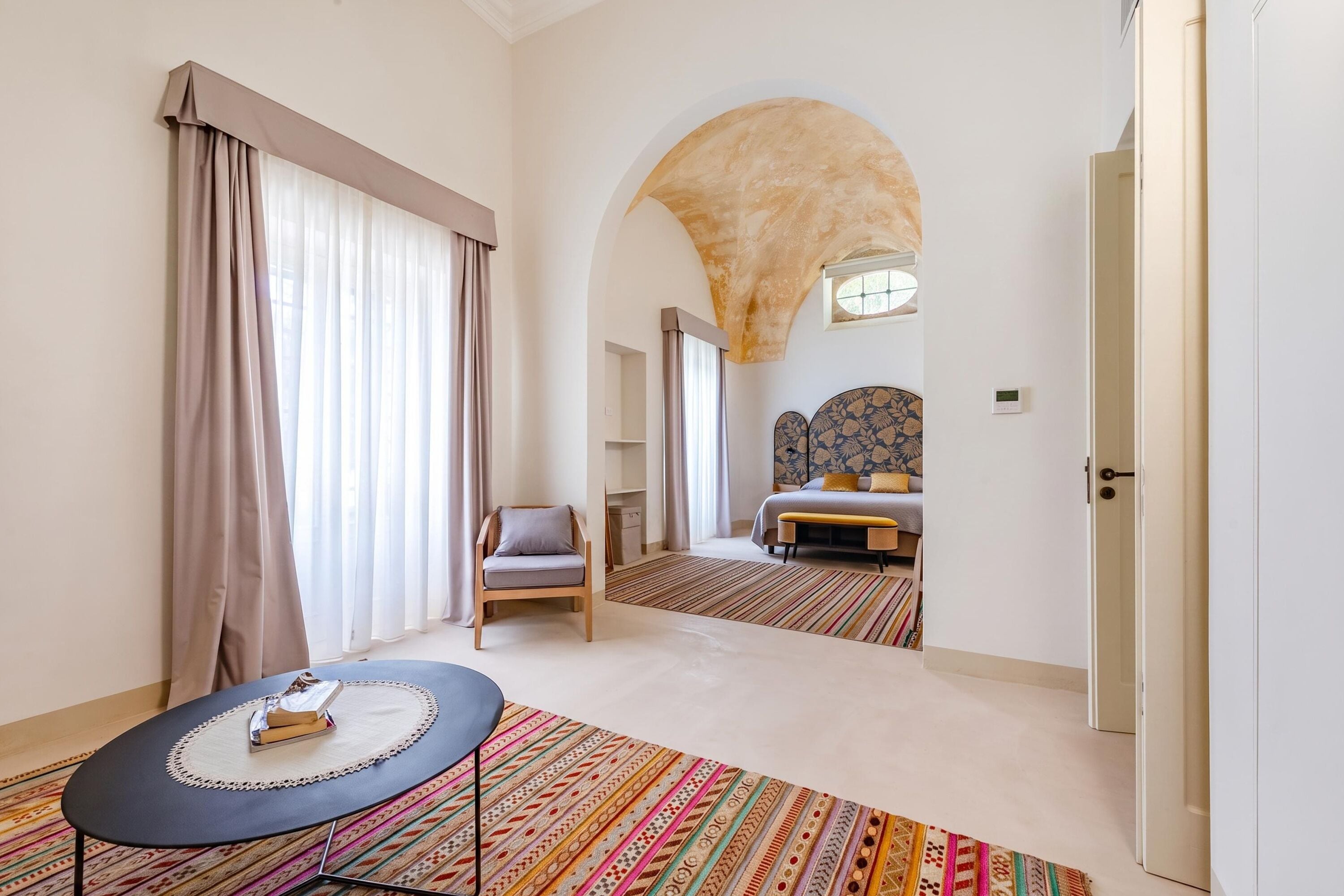 Une chambre d’hôtel avec un lit, une chaise, une table sur laquelle se trouve un livre, et un tapis coloré.
