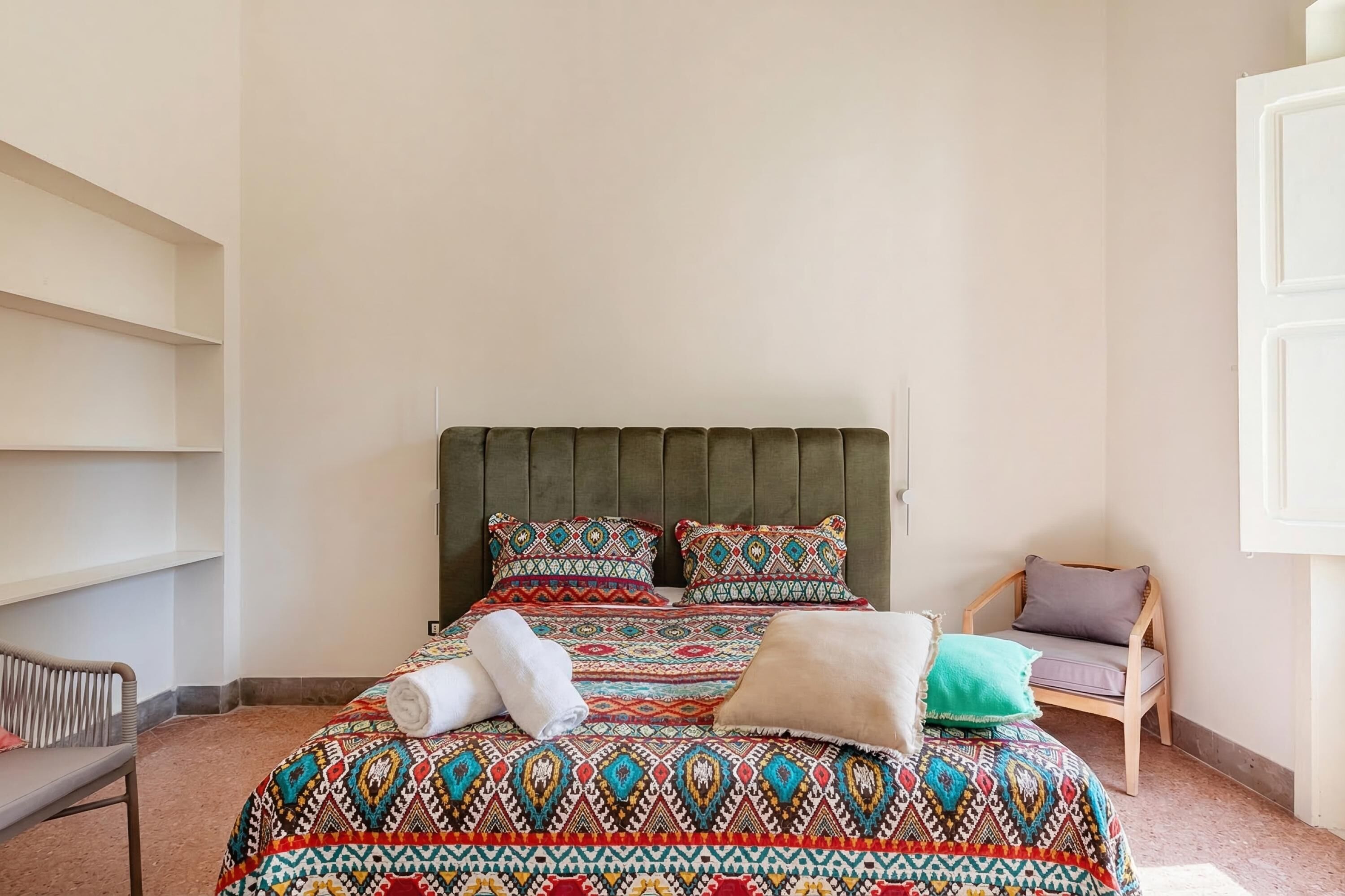 Una cama bien hecha con ropa de cama estampada, una silla de madera y una estantería en una habitación con paredes lisas.