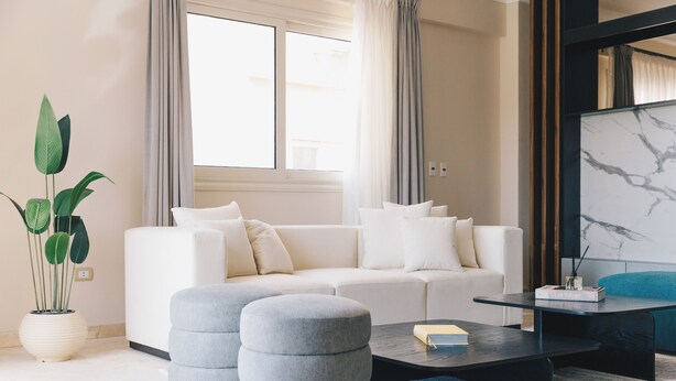 A modern living room with a white sofa, two grey ottomans, a black coffee table, and a large window with curtains. - VIDA in Yasmeen - V201, VIDA Garden View Apartment (New Cairo City)