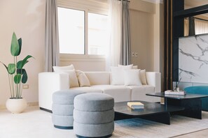 Un salon moderne avec un canapé blanc, deux poufs gris, une table basse noire et une grande fenêtre avec des rideaux.