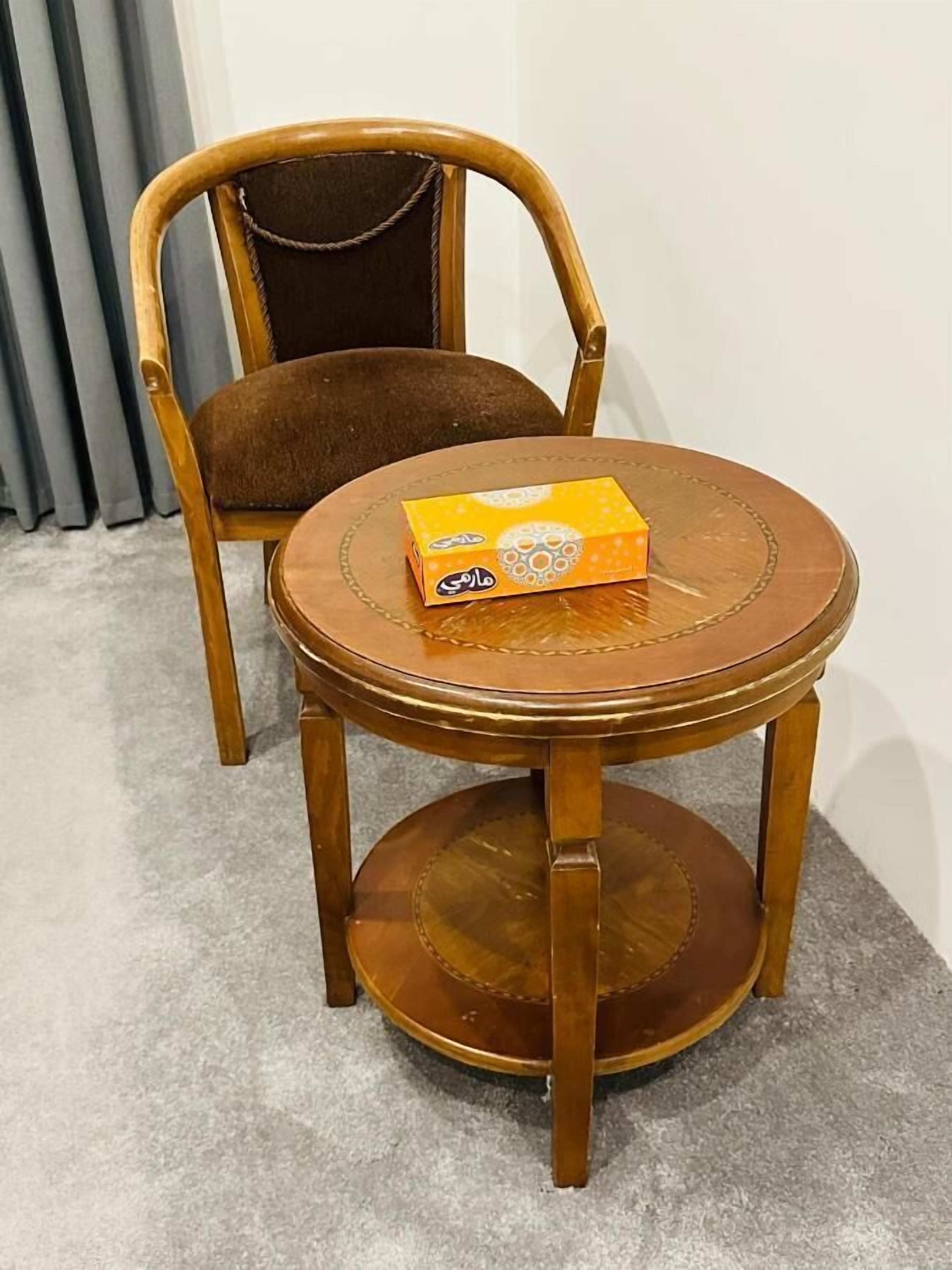 A round wooden table with a box on top, a wooden chair with a cushioned seat, and a grey carpeted floor.