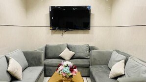 Un salon moderne avec un téléviseur à écran plat fixé au mur, deux canapés gris avec des coussins, une table basse en bois agrémentée d’un arrangement floral et un petit fauteuil.
