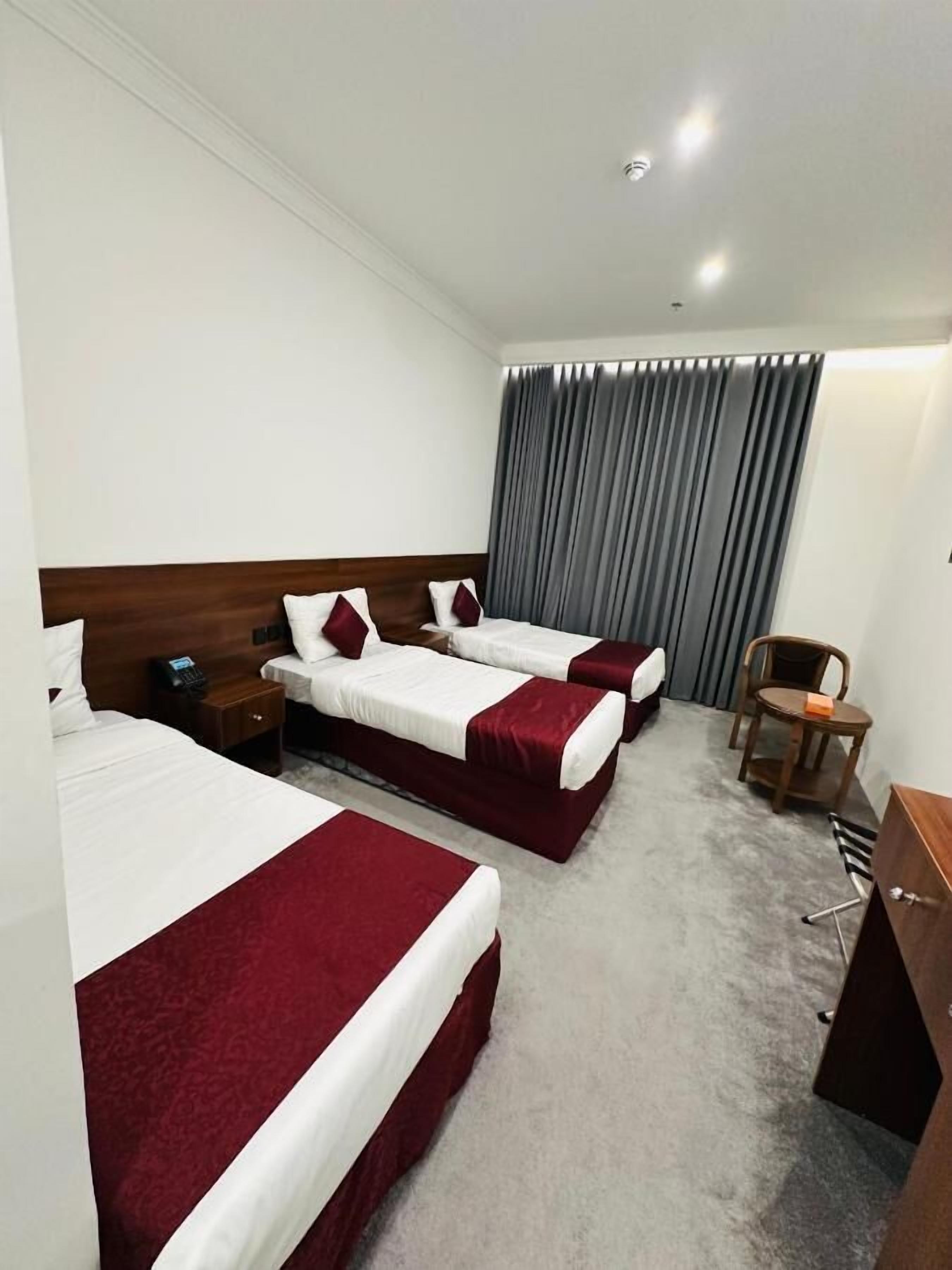 Une chambre d’hôtel avec trois lits simples, un bureau, une chaise et une grande fenêtre avec des rideaux.