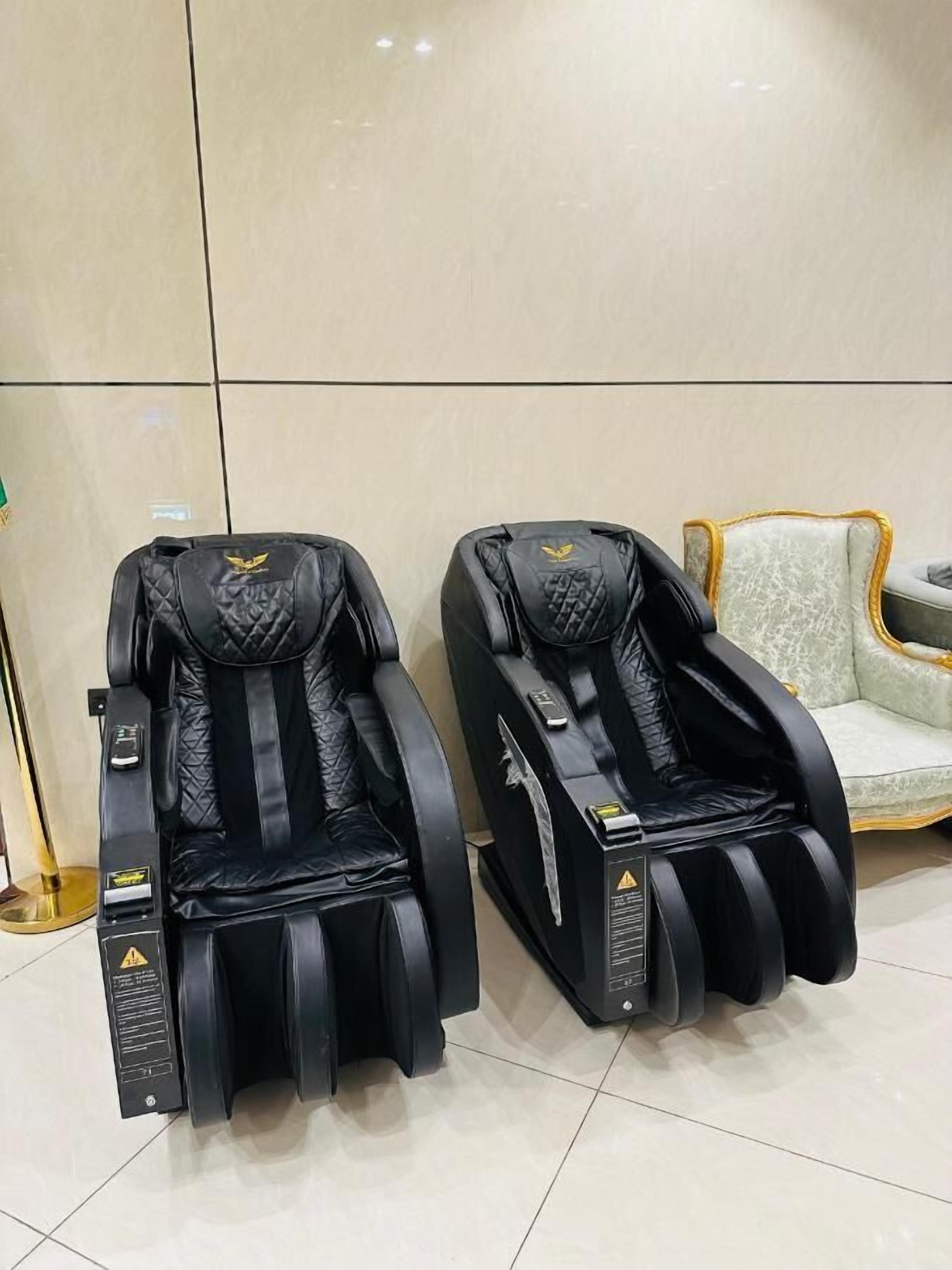 Dos sillones de masaje negros con paneles de control, ubicados en una habitación con piso de baldosas y un sofá de color claro al fondo.