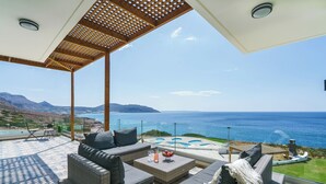 Un área de estar techada al aire libre con mobiliario de mimbre, una piscina y vista al mar.