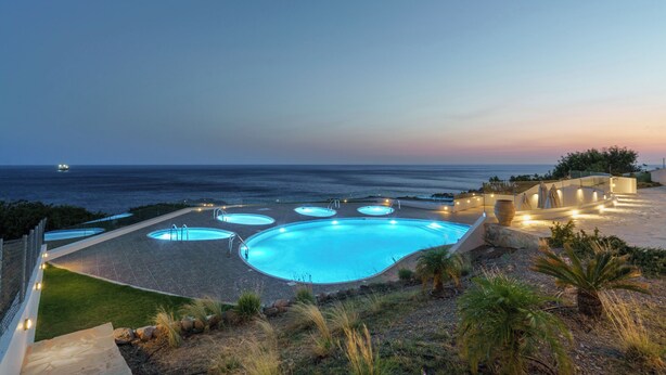 Un área de piscina con varias piscinas circulares y una piscina central grande, rodeada por un jardín ajardinado y vistas al océano.
