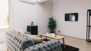 A modern living room with a tufted grey sofa, a small wooden coffee table, a potted plant, and a flat-screen TV mounted on the wall.