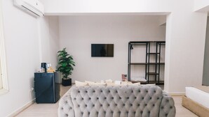 A modern living room with a grey tufted sofa, a black metal shelf unit, a potted plant, and a flat-screen TV on the wall.