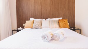 A neatly made bed with white linens and rolled towels, a wooden headboard, and a variety of patterned pillows. - VIDA in Yasmeen - V402, Vida New Standard Studio (New Cairo City)