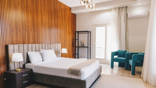 Une chambre moderne avec un grand lit, des tables de chevet, une commode, un mur d’accent en bois, une étagère avec des objets décoratifs et des fauteuils de couleur turquoise.