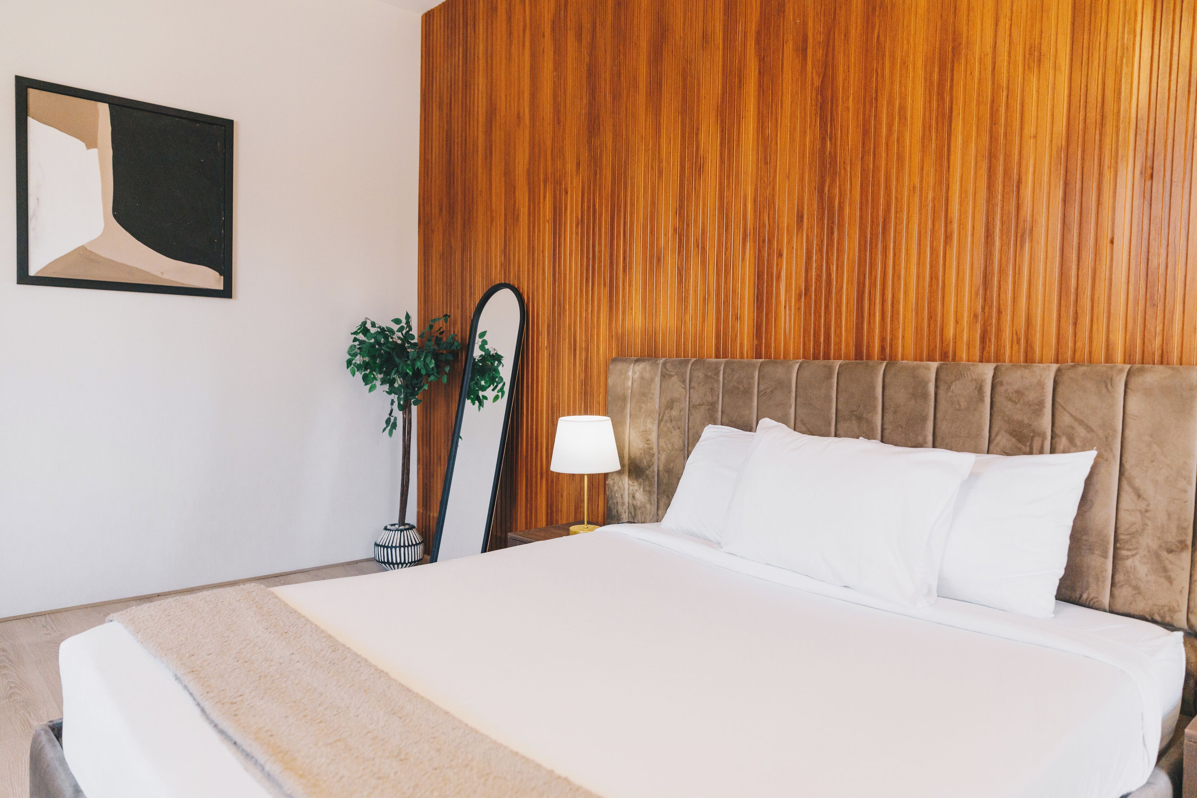 A neatly made bed with white linens and a beige throw, a bedside lamp, and a mirror on a wooden wall.