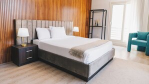 Une chambre moderne avec une tête de lit en bois, un lit recouvert de linge de maison blanc, deux tables de chevet avec des lampes et un fauteuil turquoise.