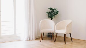 Une pièce avec deux chaises blanches, une plante en pot et une grande fenêtre avec des rideaux blancs.