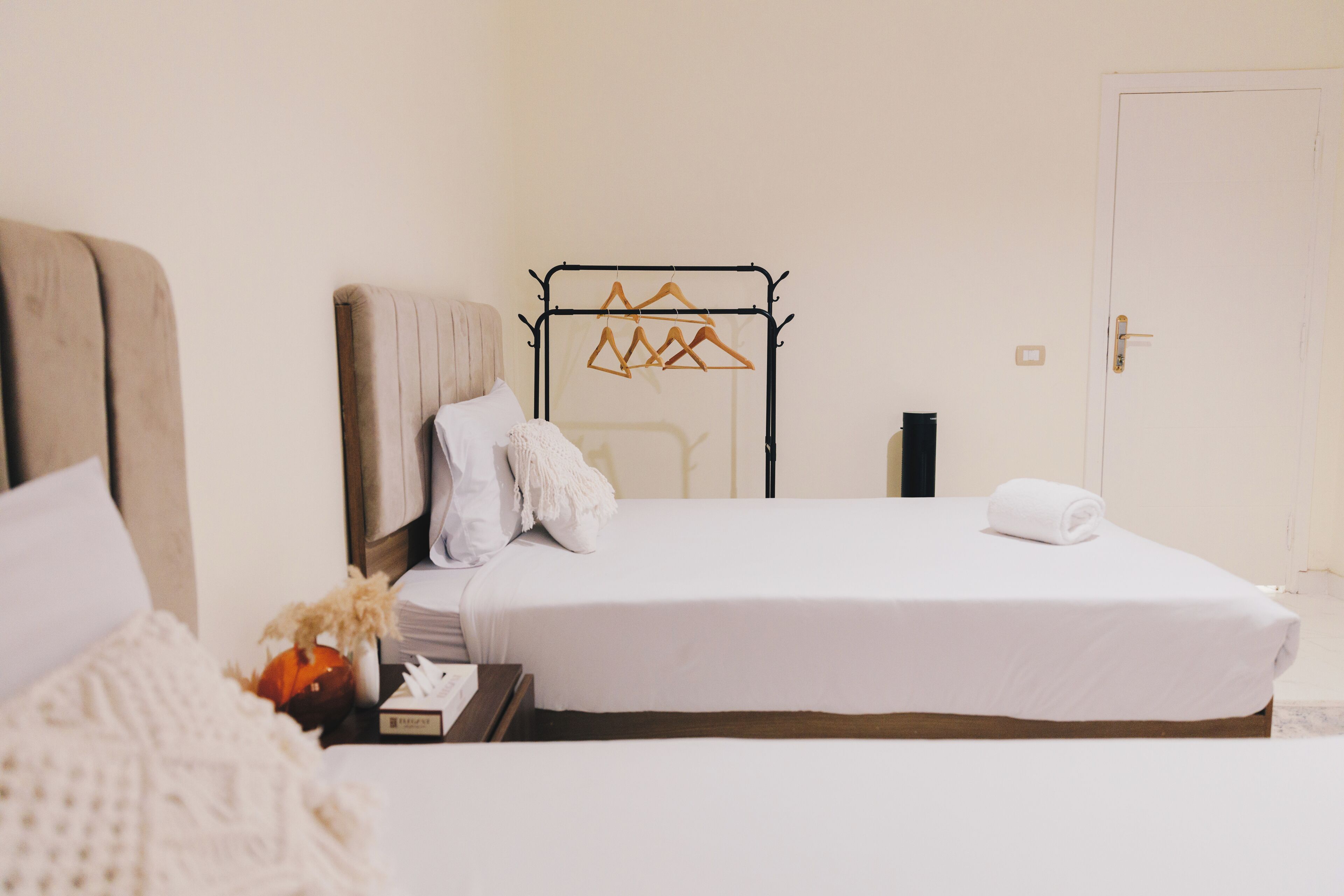 Una cama bien hecha con ropa de cama blanca, una mesita de noche con una lámpara y un perchero metálico con perchas.