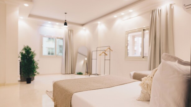 A modern bedroom with a large bed, a bedside table, a hanging light fixture, a window with curtains, and a potted plant. - VIOLA Near Waterway - 21, VIOLA New Studio - Higher Floor (New Cairo City)