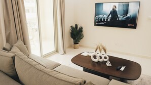 A modern living room with a large window, a sofa, a coffee table with decorative items, and a TV displaying a Netflix show. - VIOLA Near Waterway - 21, VIOLA New Studio - Higher Floor (New Cairo City)