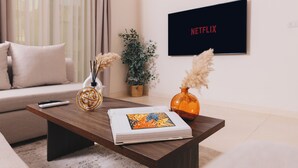 A modern living room with a sofa, a coffee table, a book, a vase, and a wall-mounted TV displaying the Netflix logo.