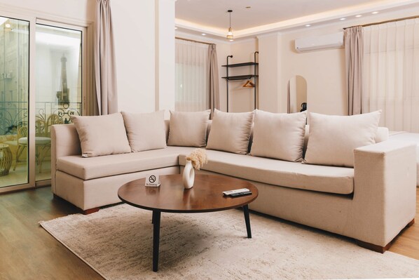 A modern living room with a beige sectional sofa, a round coffee table, and a glass door leading to a balcony. - VIOLA Near Waterway - 32, VIOLA New Studio - Higher Floor (New Cairo City)