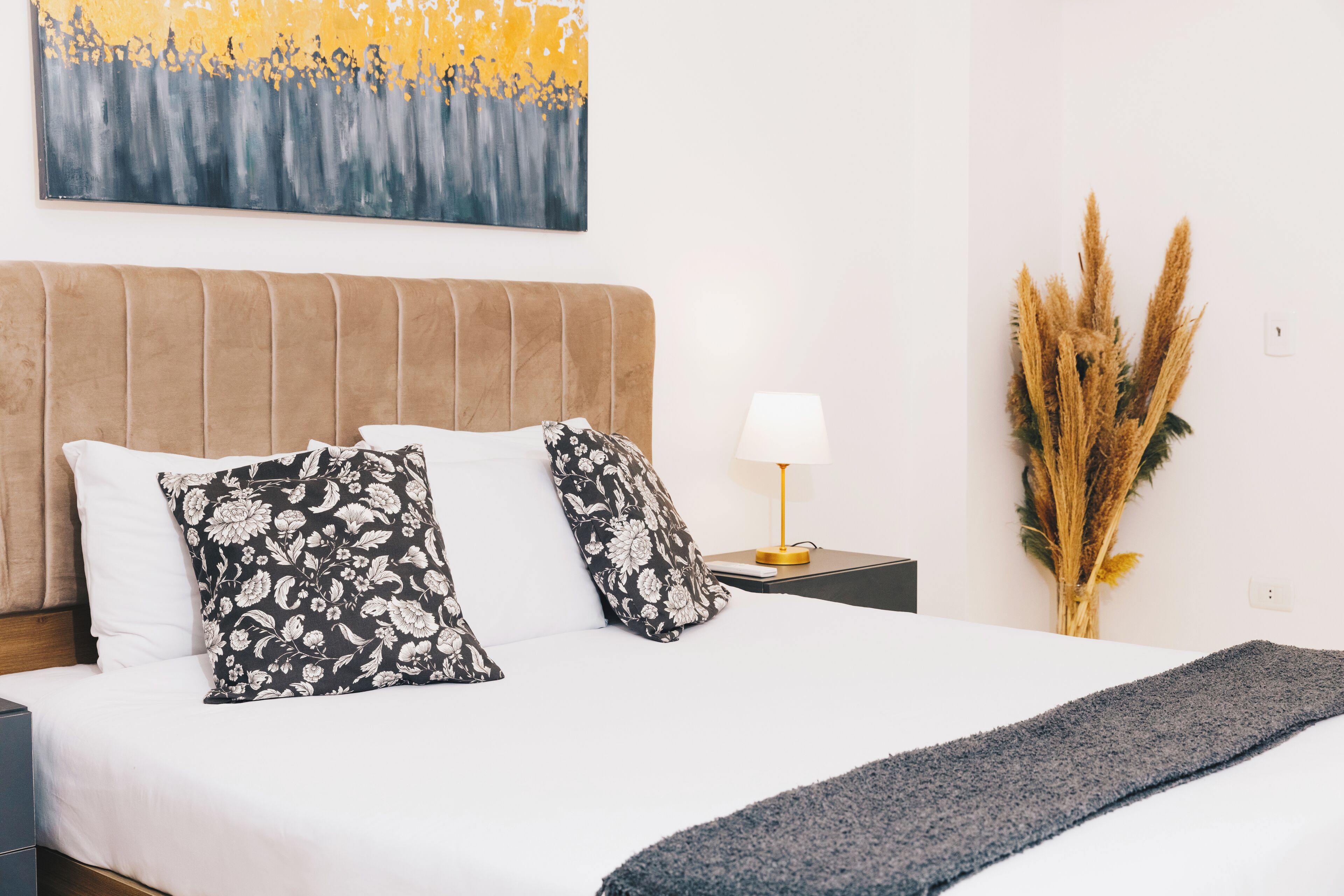 A neatly made bed with white and black floral pillows, a beige headboard, a bedside lamp, and a vase with dried plants.
