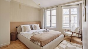 A modern bedroom with a large bed, a wooden side table, a chair, and a window with curtains. - Amazing Apartment Le Marais Center of Paris (Paris)