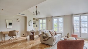 A spacious living room with a herringbone wooden floor, a sofa, a pink armchair, a dining area with a table and chairs, and large windows offering a view of buildings outside. - Amazing Apartment Le Marais Center of Paris (Paris)