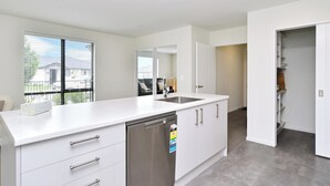 A modern kitchen with white cabinetry, a stainless steel dishwasher, and a large window providing a view of the outside. - Woodend Getaway, Modern 3-Bedroom, 2-Bathroom Home,  (Woodend)
