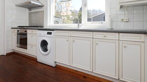 A kitchen with white cabinetry, a built-in oven, a washing machine, and a window with a view of a house and trees. - The Seahorse Bungalow Park Sollasi (Noordwijkerhout)