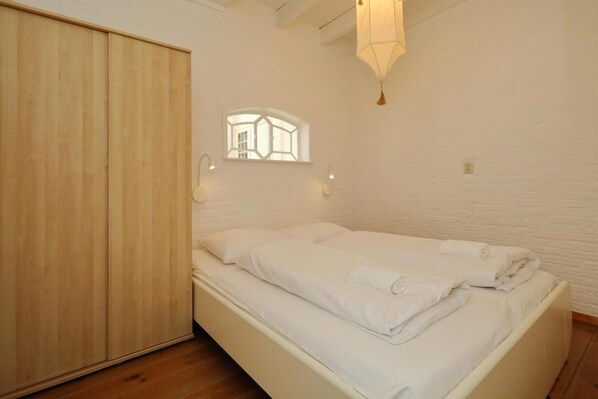 Un dormitorio con cama, armario de madera, paredes blancas, un ventanal pequeño y una lámpara colgante.