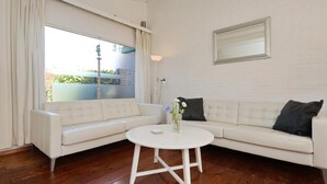 A white leather sofa with a round white coffee table in the center, a white wall with a framed mirror, and a window with curtains. - The Seahorse Bungalow Park Sollasi (Noordwijkerhout)