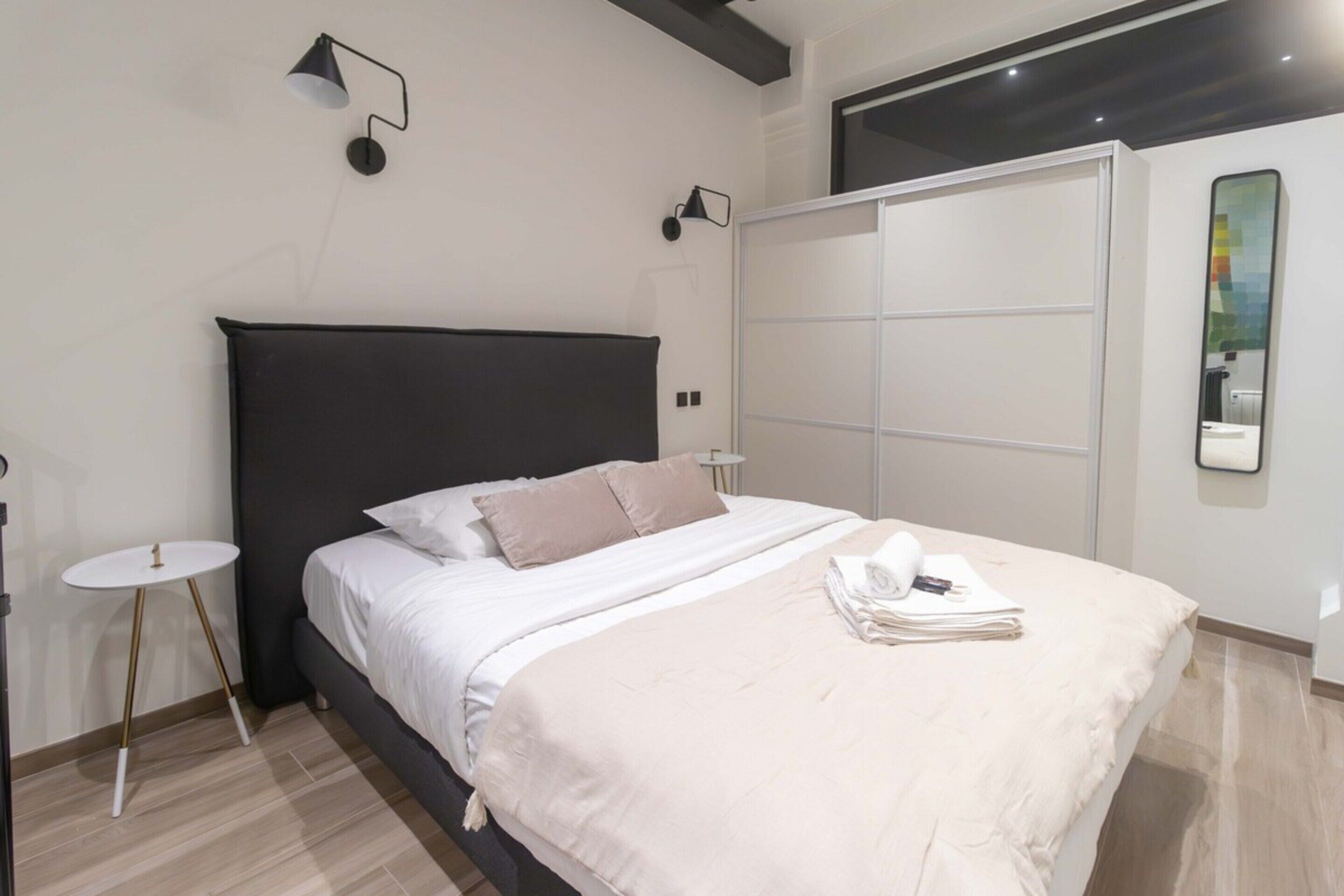 A modern bedroom with a black headboard, white bedding, a small round table, and a large mirror on the wall.