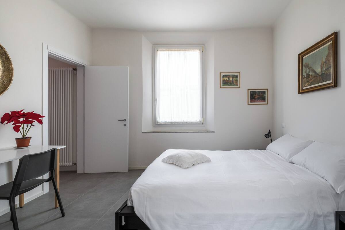 A bedroom with a bed, a desk, a chair, a radiator, a window, and a door.