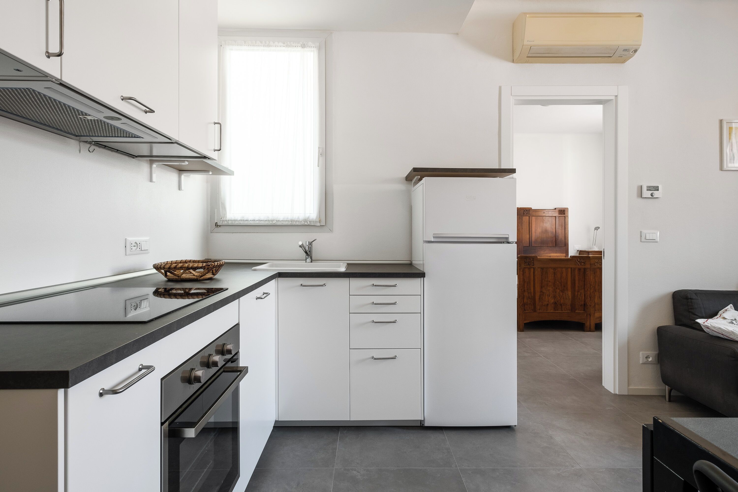 A modern kitchen with white cabinets, a black countertop, and a built-in oven. There is a window providing natural light and a view into the dining area.