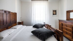 Una camera da letto con testiera in legno, un letto con lenzuola bianche, due cuscini neri, un comodino con una lampada, una cassettiera in legno e un quadro incorniciato appeso al muro.
