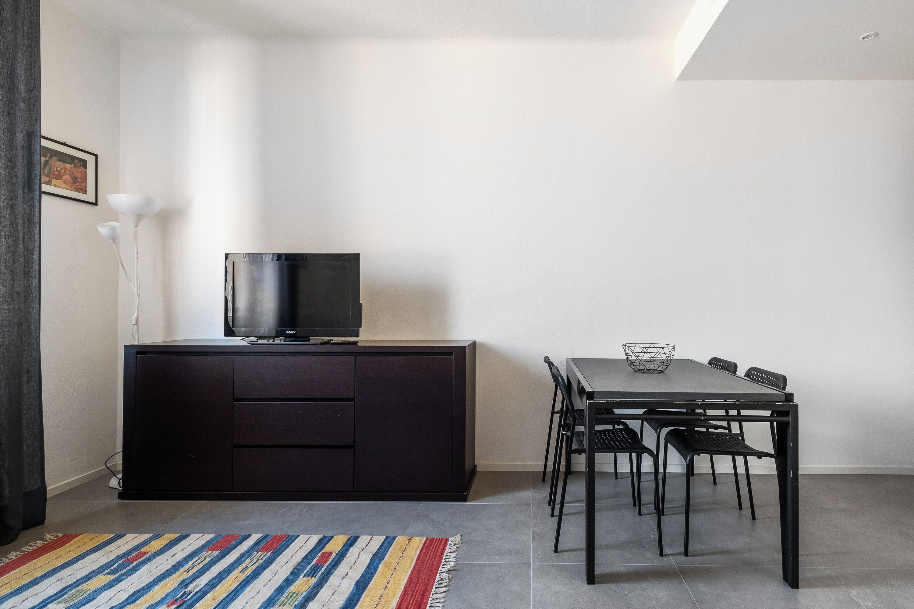 A modern room with a dark wooden cabinet, a flat-screen TV, a dining table with chairs, and a colorful striped rug.