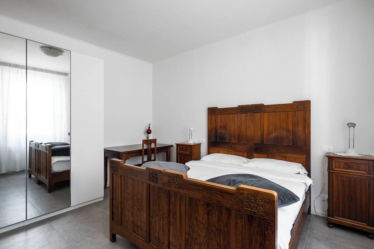 A bedroom with a wooden bed, a desk, a chair, a wardrobe, and a mirror.