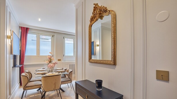 Comedor con una mesa lista para cuatro, un espejo grande y ornamentado, y una consola con un jarrón de flores.