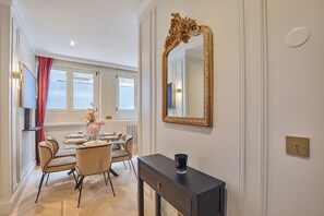 Uma área de refeições com uma mesa posta para quatro pessoas, um espelho grande e decorativo, e um aparador com um vaso de flores.