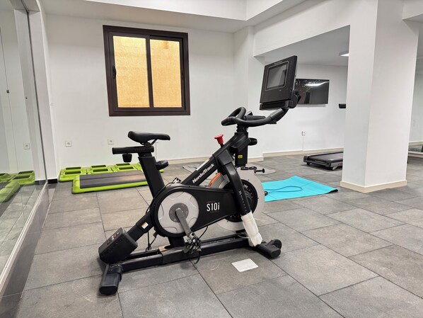 Una bicicleta estática, una caminadora y colchonetas de ejercicio en una habitación con paredes blancas y un ventanal grande.