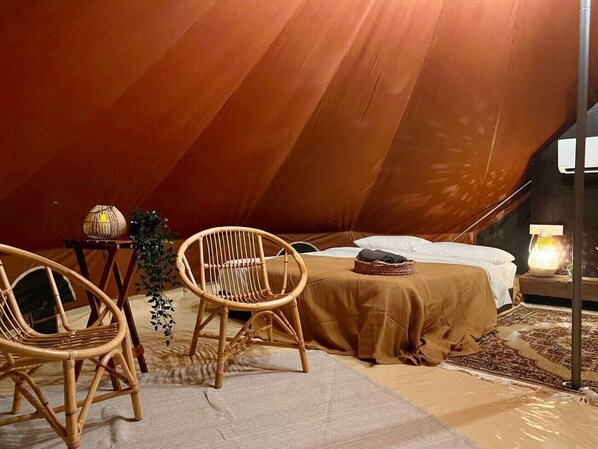 Een gezellig ingerichte tent met een bed, twee rieten stoelen, een kleine tafel met een lamp en een decoratieve plant.